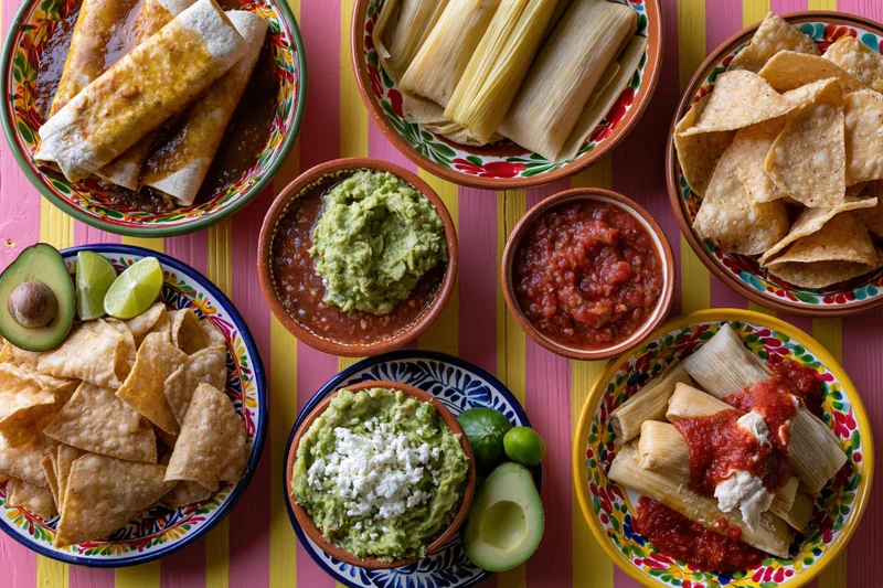 Overhead flat-lay of a traditional mexican food spread with enchiladas, tamales, guacamole, salsa, chips, and avocado on a colorful striped table
