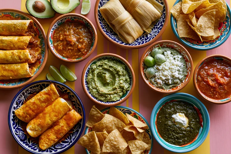 Overhead flat-lay of a traditional mexican food spread with enchiladas, tamales, guacamole, salsa, chips and lime on a colorful table