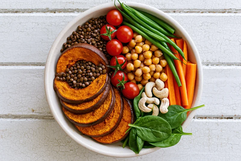 Overhead view of a ceramic bowl filled with lentils, chickpeas, sweet potato, carrots, green beans, tomatoes, spinach and cashews on a white wooden surface