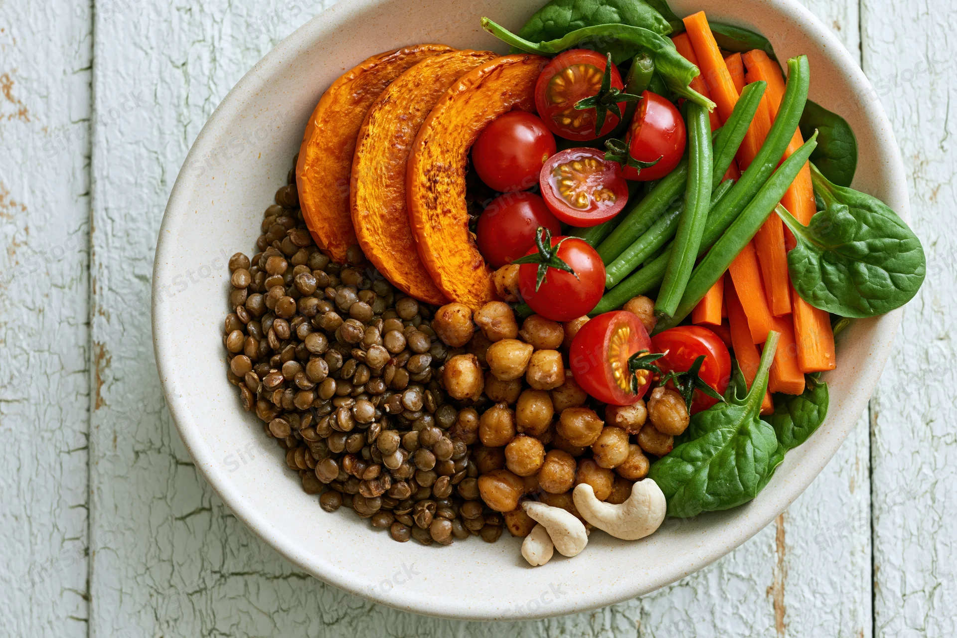 Overhead view of a ceramic bowl filled with lentils, chickpeas, roasted squash, cherry tomatoes, green beans, carrots, spinach and cashews on a white wooden surface