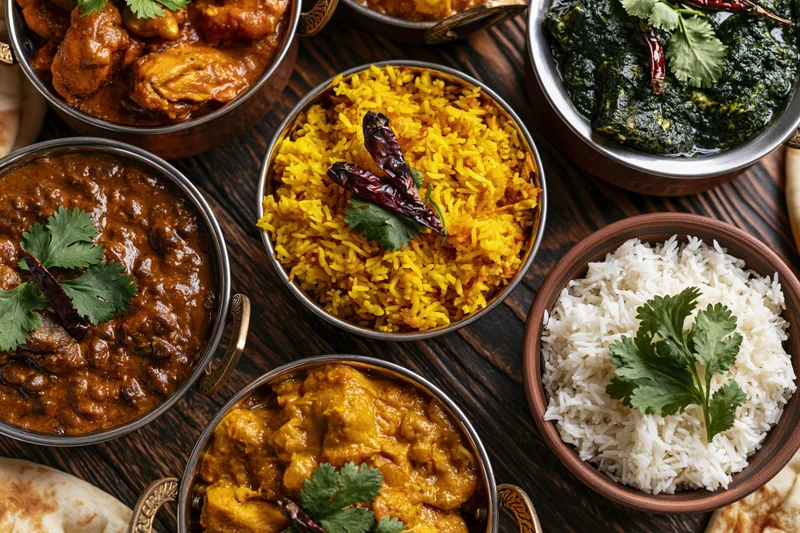 Overhead flat-lay of an indian feast with curries, rice, dal, and naan on a dark wooden table