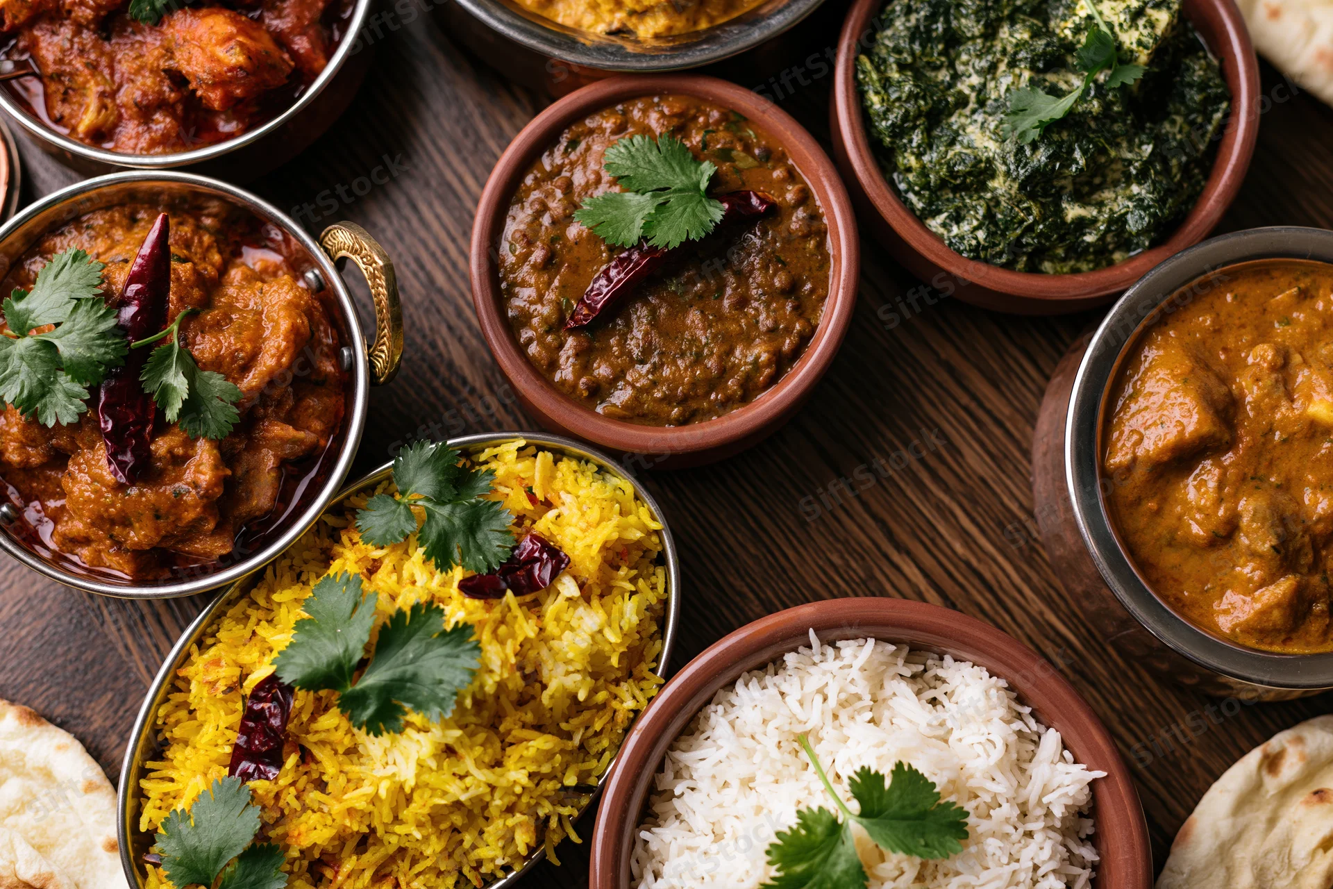 Overhead flat-lay of an indian feast with curries, rice, naan and garnishes on a dark wooden table