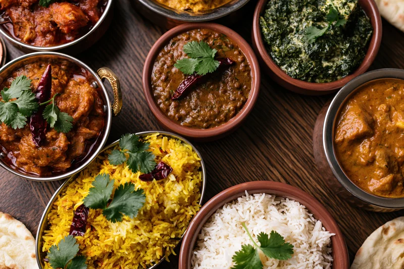 Overhead flat-lay of an indian feast with curries, rice, naan and garnishes on a dark wooden table