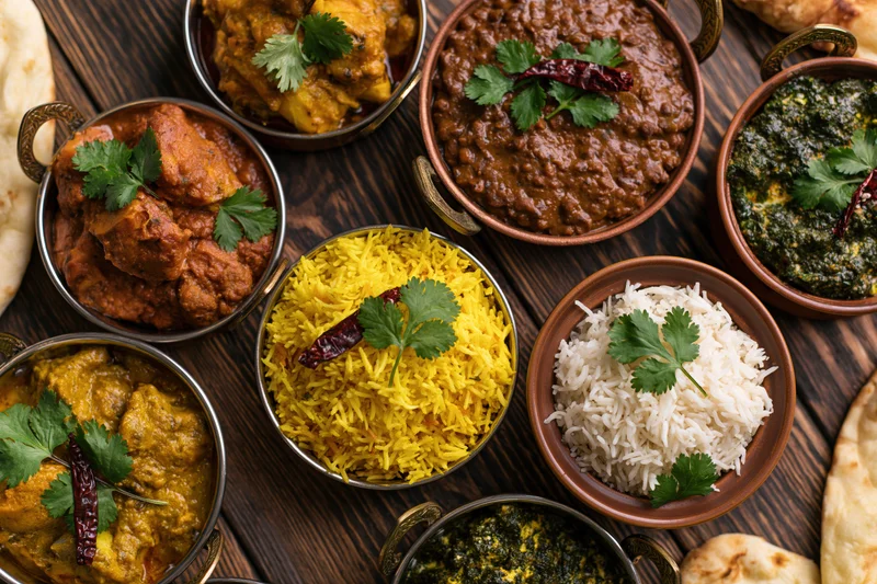 Indian feast spread with curries, rice, naan and garnishes on dark wooden table, overhead flat-lay view