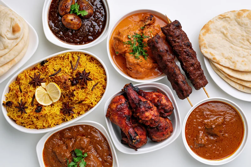 Overhead flat-lay of an indian nonvegetarian feast with biryani, curries, kebabs, tandoori chicken, and naan bread arranged on white plates