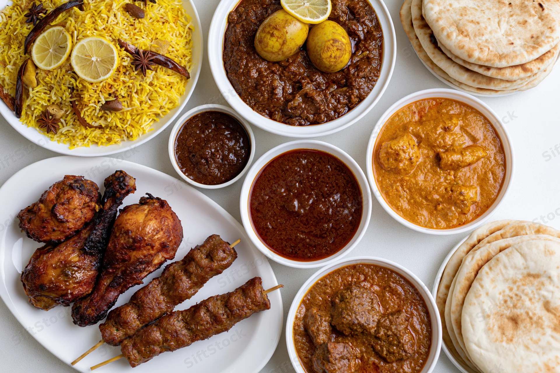 Overhead flat-lay of an indian nonvegetarian feast with biryani, curries, tandoori chicken, kebabs, and naan bread arranged on white plates and bowls