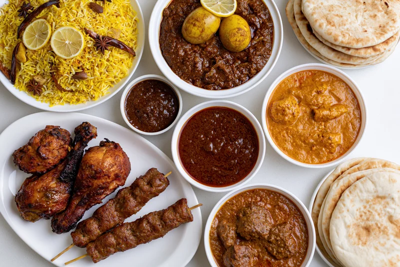 Overhead flat-lay of an indian nonvegetarian feast with biryani, curries, tandoori chicken, kebabs, and naan bread arranged on white plates and bowls