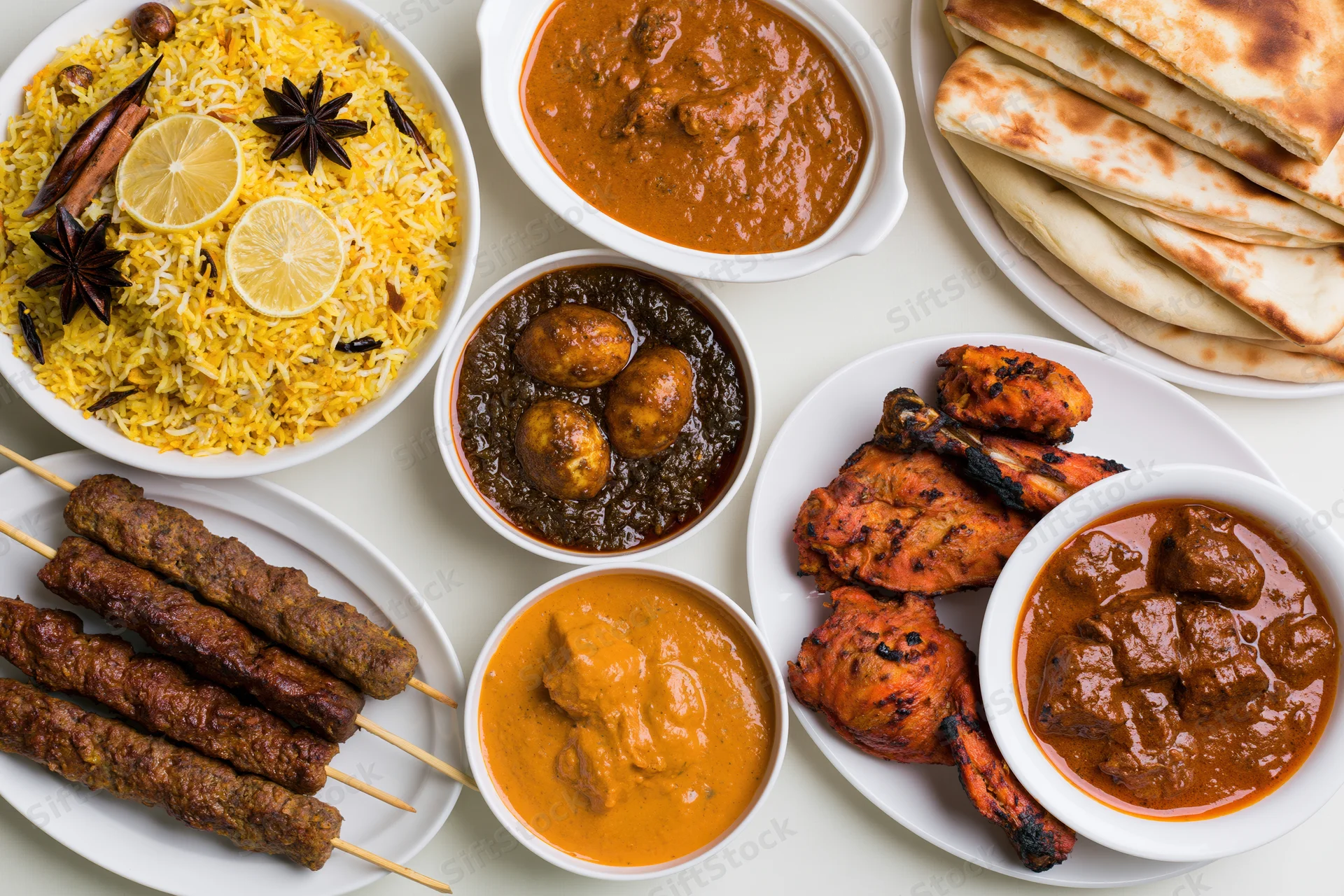 Overhead flat-lay of an indian nonvegetarian feast spread with biryani, curries, kebabs, tandoori chicken, and naan on white plates