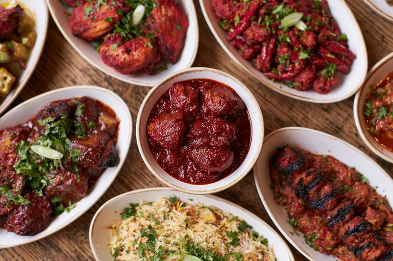 Overhead view of an indian restaurant meal spread on a wooden table with multiple dishes including curry, rice, kebabs, and garnishes