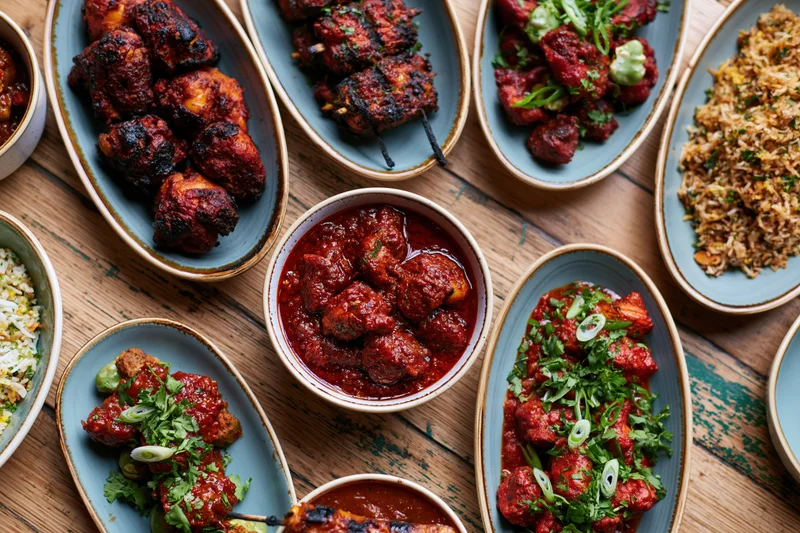 Overhead view of an indian restaurant meal spread on a wooden table with various dishes including tandoori chicken, kebabs, biryani, and curries served in blue ceramic bowls and plates