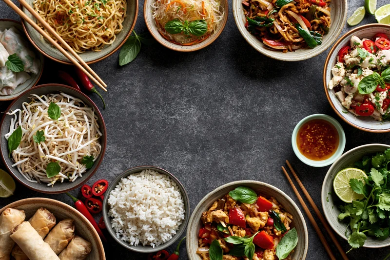 Assorted asian dishes arranged in a circular flat lay on dark textured surface with chopsticks and garnishes