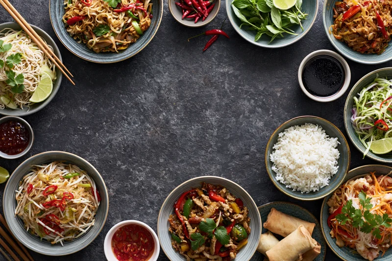Assorted asian dishes arranged in a circular flat lay on dark stone surface with copyspace in center