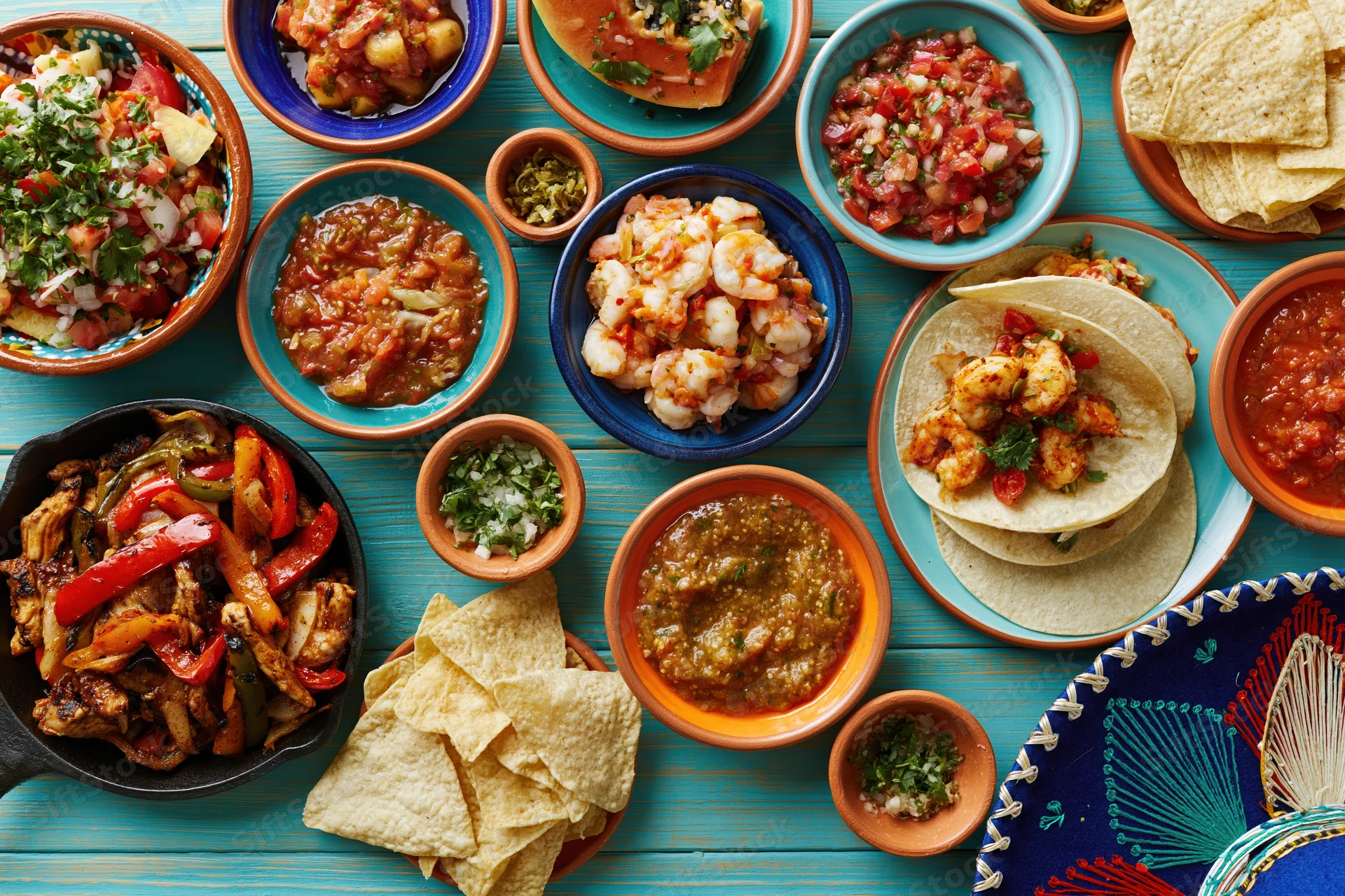 Overhead flat-lay of assorted traditional Mexican dishes on a blue wooden table with tortillas, tacos, salsa, and chips arranged around a decorative textile