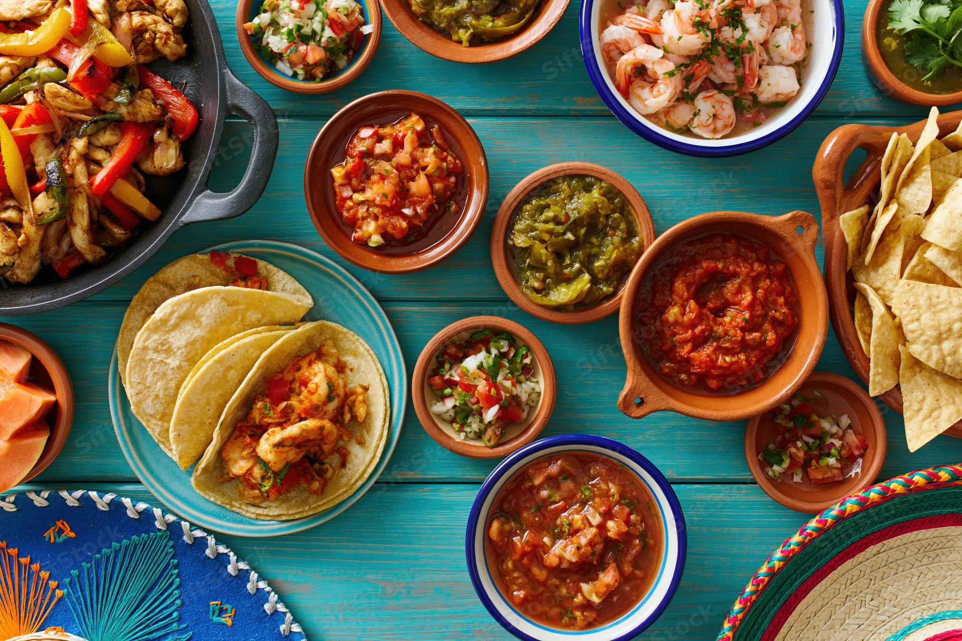 Overhead flat-lay of assorted traditional mexican dishes on a blue wooden table including tacos, salsa, guacamole, shrimp, and chips