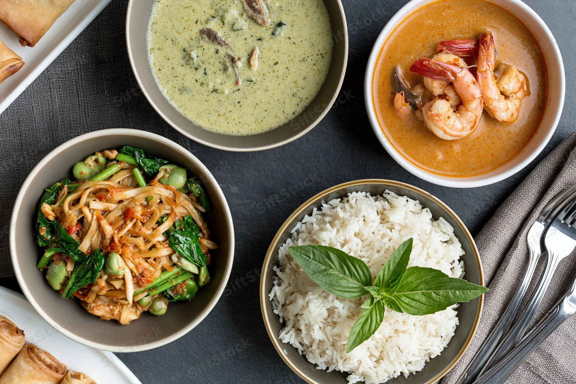 Overhead flat-lay of four thai dishes including curry, rice, noodles and spring rolls arranged on dark slate table