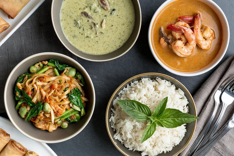 Overhead flat-lay of four thai dishes including curry, rice, noodles and spring rolls arranged on dark slate table