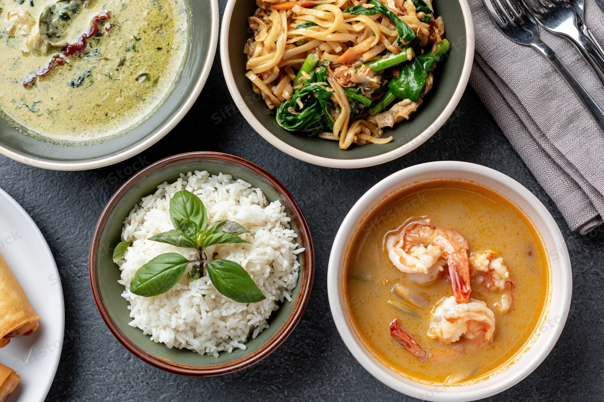 Overhead flat-lay of four thai dishes arranged on dark slate surface including curry, noodles, rice, and shrimp soup