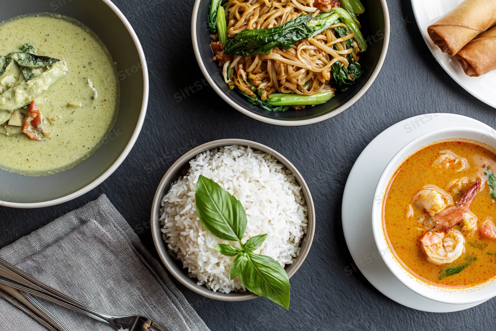 Overhead flat-lay of four thai dishes arranged on dark slate table with bowls of curry, noodles, rice, and spring rolls
