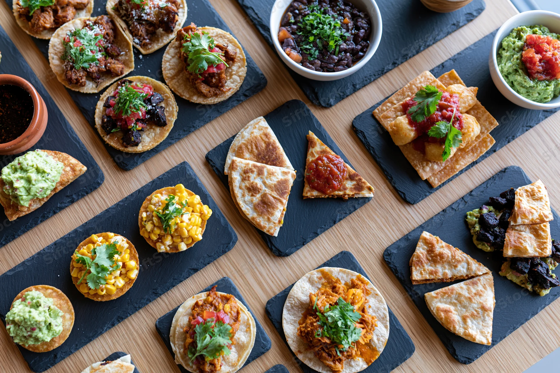 Overhead flat-lay of multiple mexican restaurant dishes arranged on slate boards and wooden table
