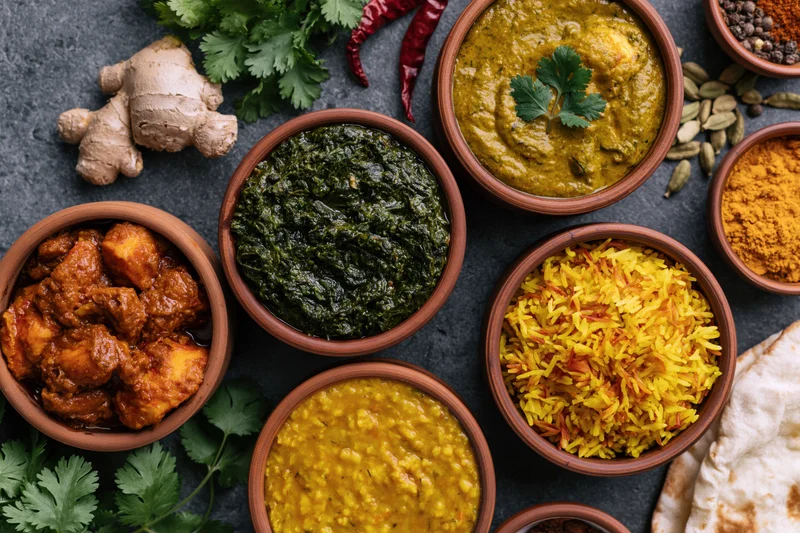 Overhead flat-lay of indian dishes in clay bowls arranged on dark stone surface with spices and herbs