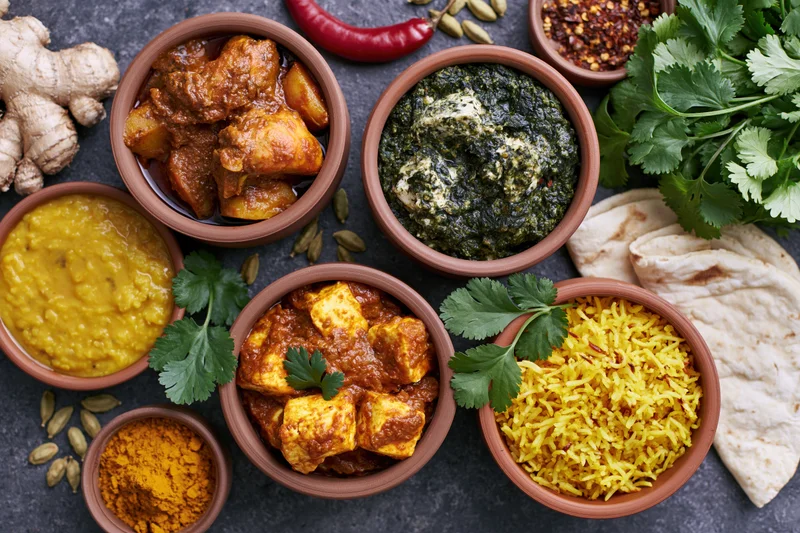 Overhead flat-lay of indian dishes in clay bowls with curry, rice, naan, and spices arranged on dark surface