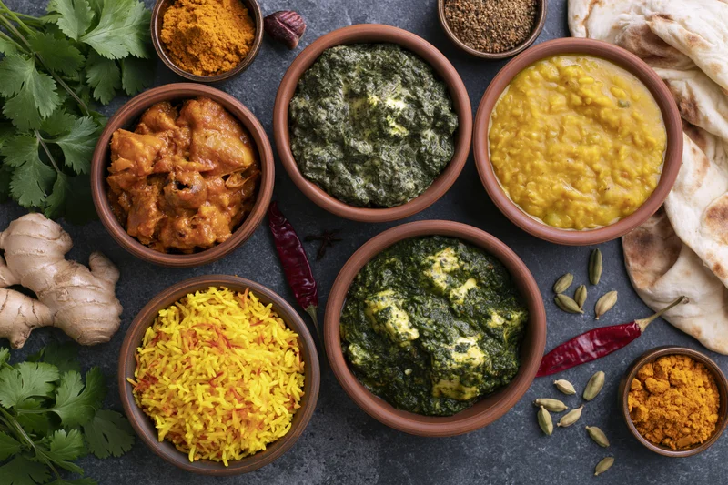 Overhead flat-lay of indian dishes in clay bowls with spices and naan on dark stone surface