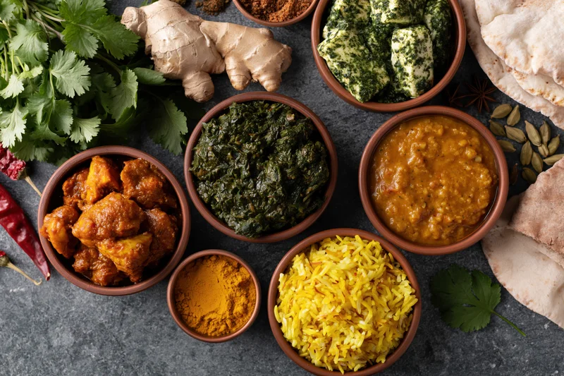 Overhead flat-lay of indian dishes in clay bowls arranged on dark stone surface with spices and herbs