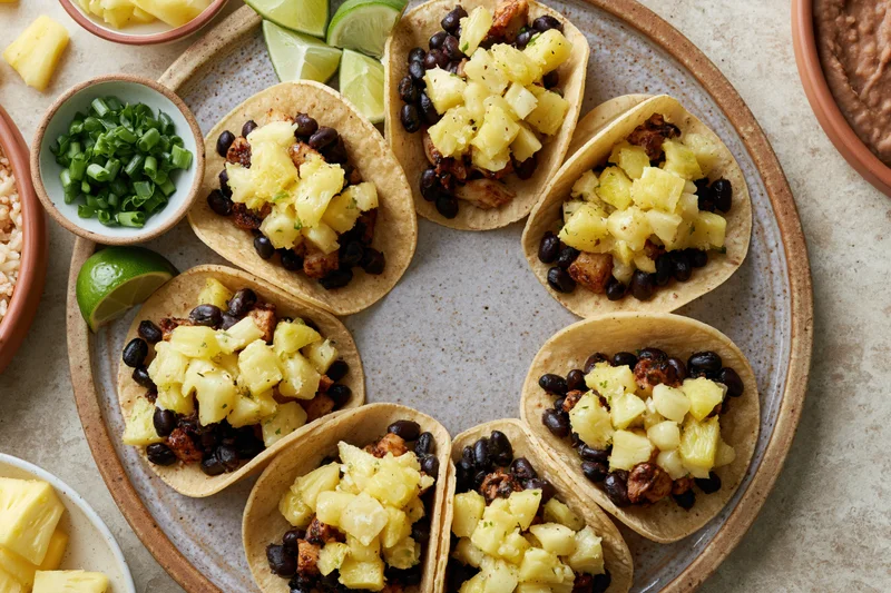 Six small corn tortilla tacos topped with pineapple and black beans arranged on a ceramic plate with lime and green onions nearby