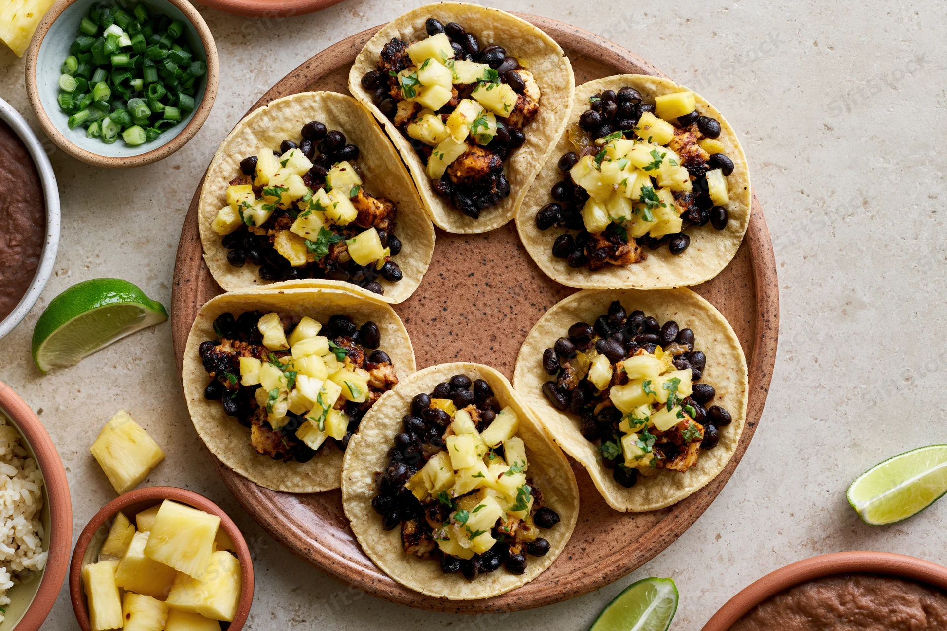 Six small corn tortilla tacos topped with black beans, pineapple, and cilantro arranged on a ceramic plate with lime wedges and side bowls of rice and refried beans on a countertop