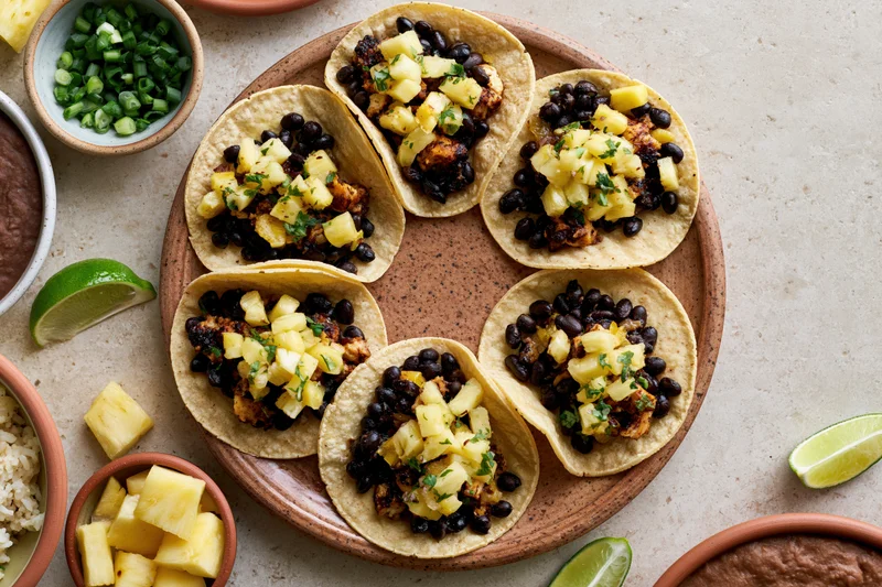 Six small corn tortilla tacos topped with black beans, pineapple, and cilantro arranged on a ceramic plate with lime wedges and side bowls of rice and refried beans on a countertop