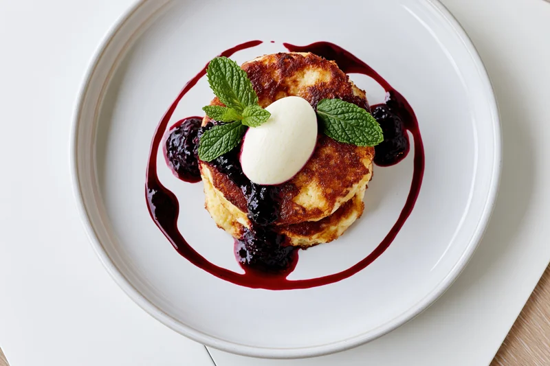 Stacked golden cheese pancakes with berry sauce and mint garnish on white plate
