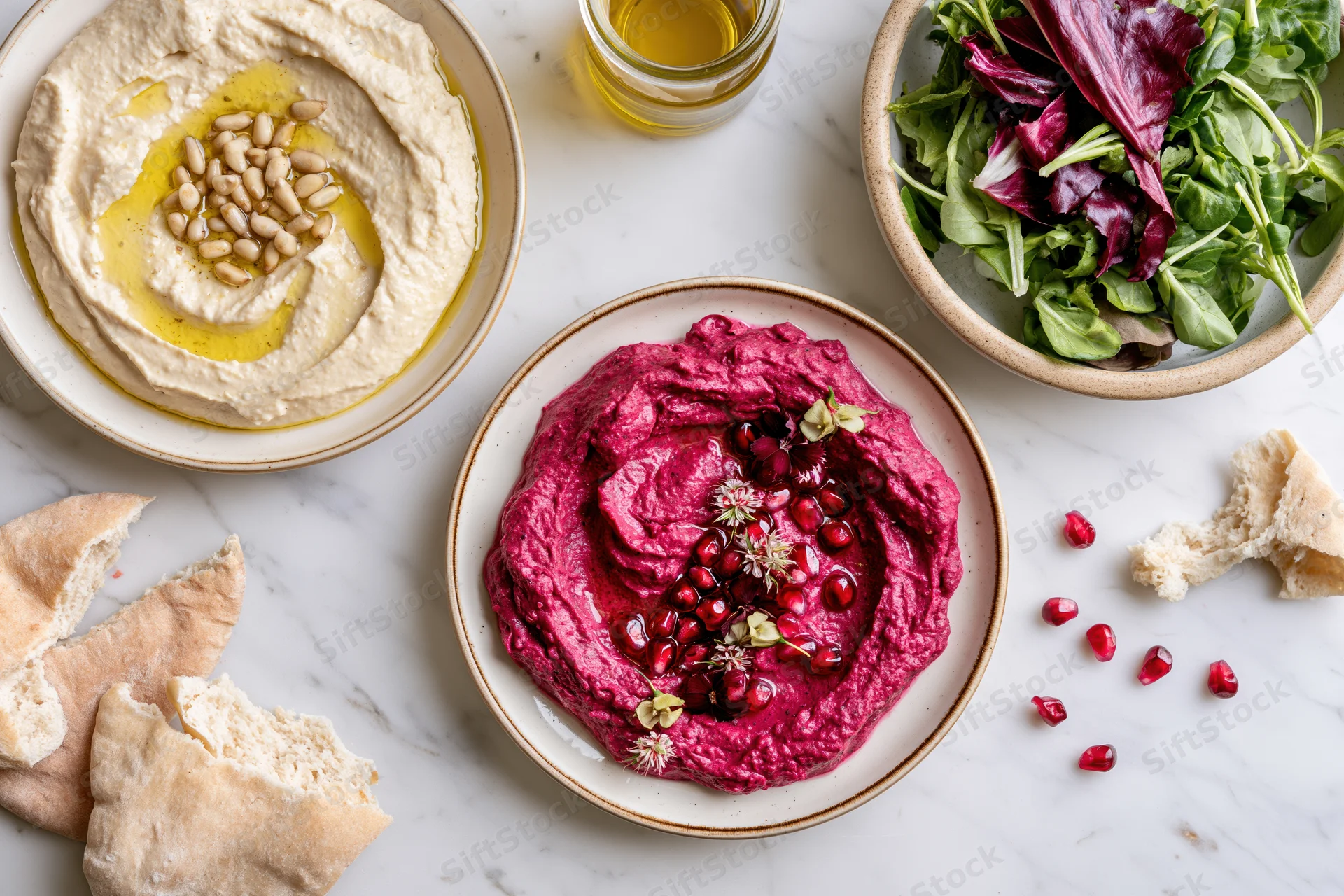 Overhead flat-lay of hummus, beetroot dip and salad on marble with pita bread and olive oil