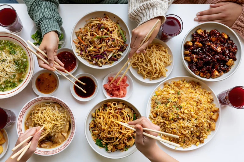 Overhead view of hands using chopsticks to eat various asian dishes from shared bowls on a white table