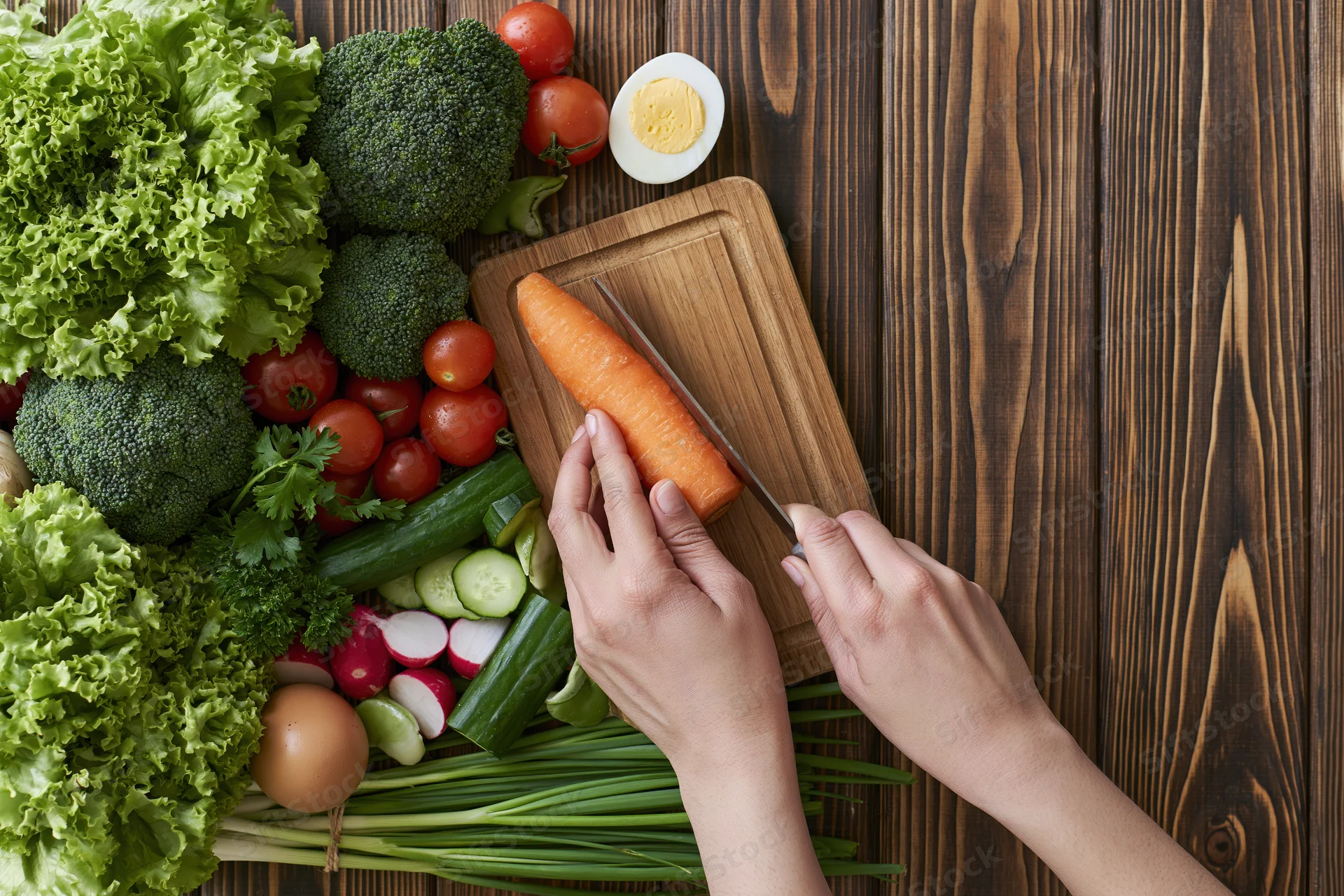 Hands slicing a carrot on a wooden cutting board surrounded by fresh vegetables on a dark wood table