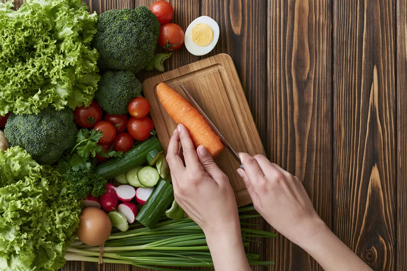 Hands slicing a carrot on a wooden cutting board surrounded by fresh vegetables on a dark wood table