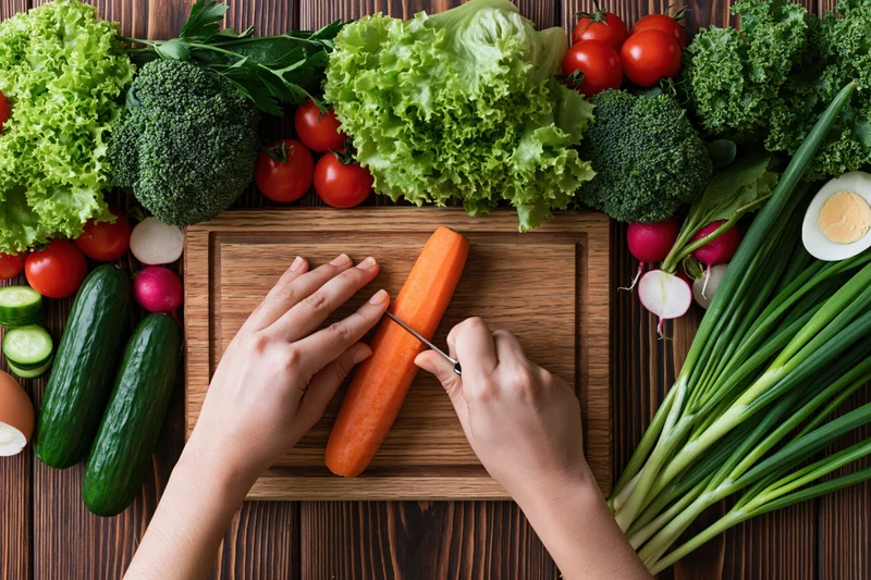 Hands slicing a carrot on a wooden cutting board surrounded by fresh vegetables