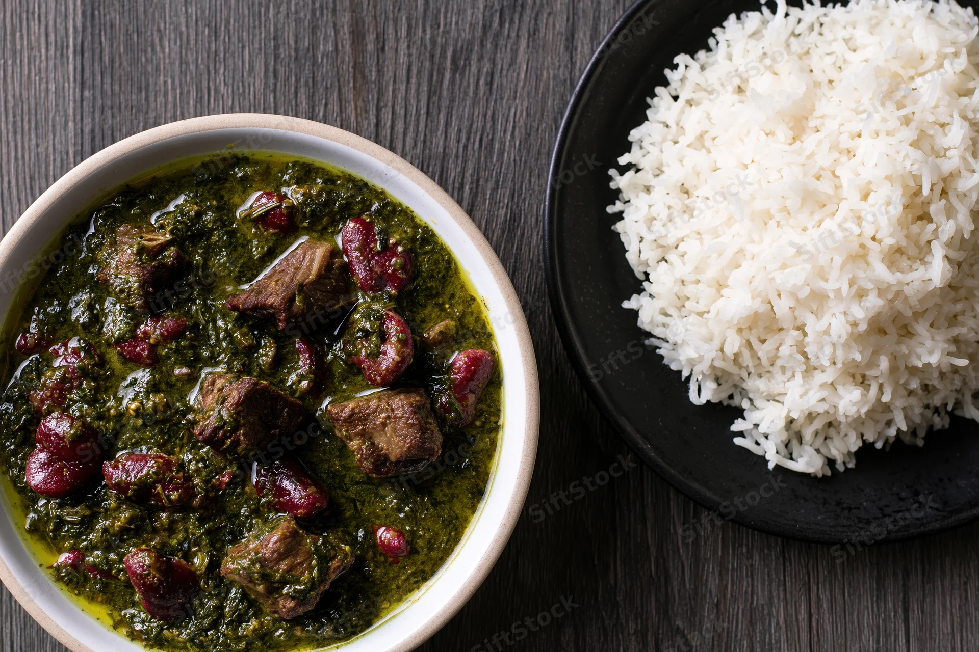 Green curry with beef and red kidney beans served in a white bowl beside a black plate of white rice on a dark wood table