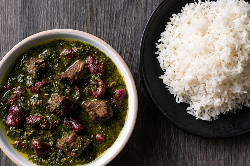 Green curry with beef and red kidney beans served in a white bowl beside a black plate of white rice on a dark wood table
