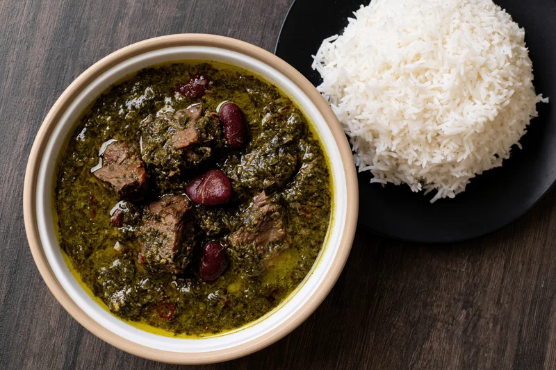 Overhead view of a bowl of green beef curry with kidney beans served next to a plate of white rice on a dark wood table