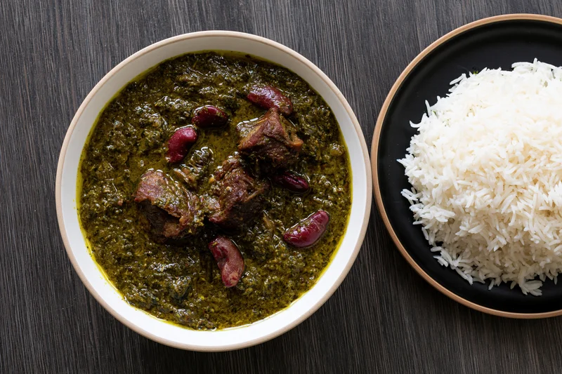 Overhead view of a bowl of green beef curry with kidney beans next to a plate of white rice on a dark wood table