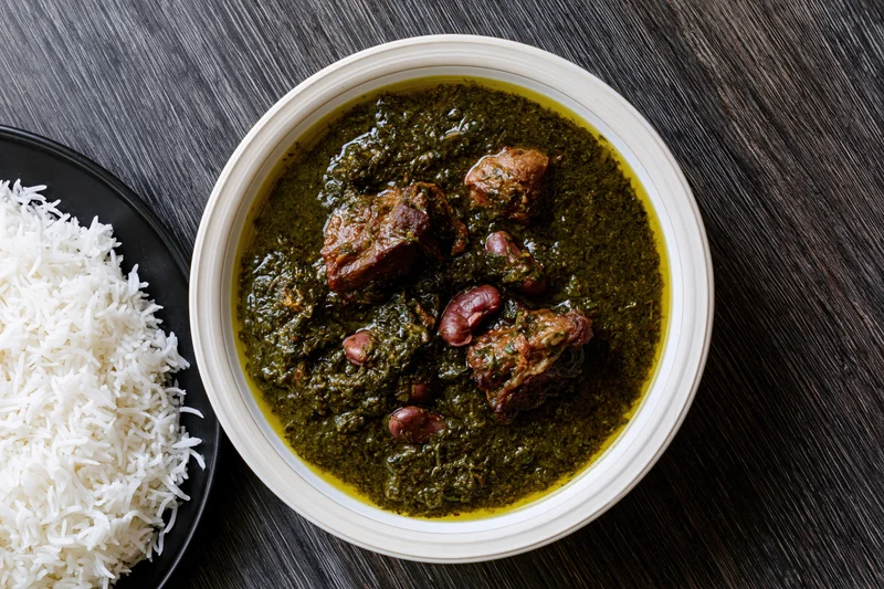Overhead view of a bowl of green spinach curry with beef and kidney beans served alongside a plate of white rice on a dark wooden table