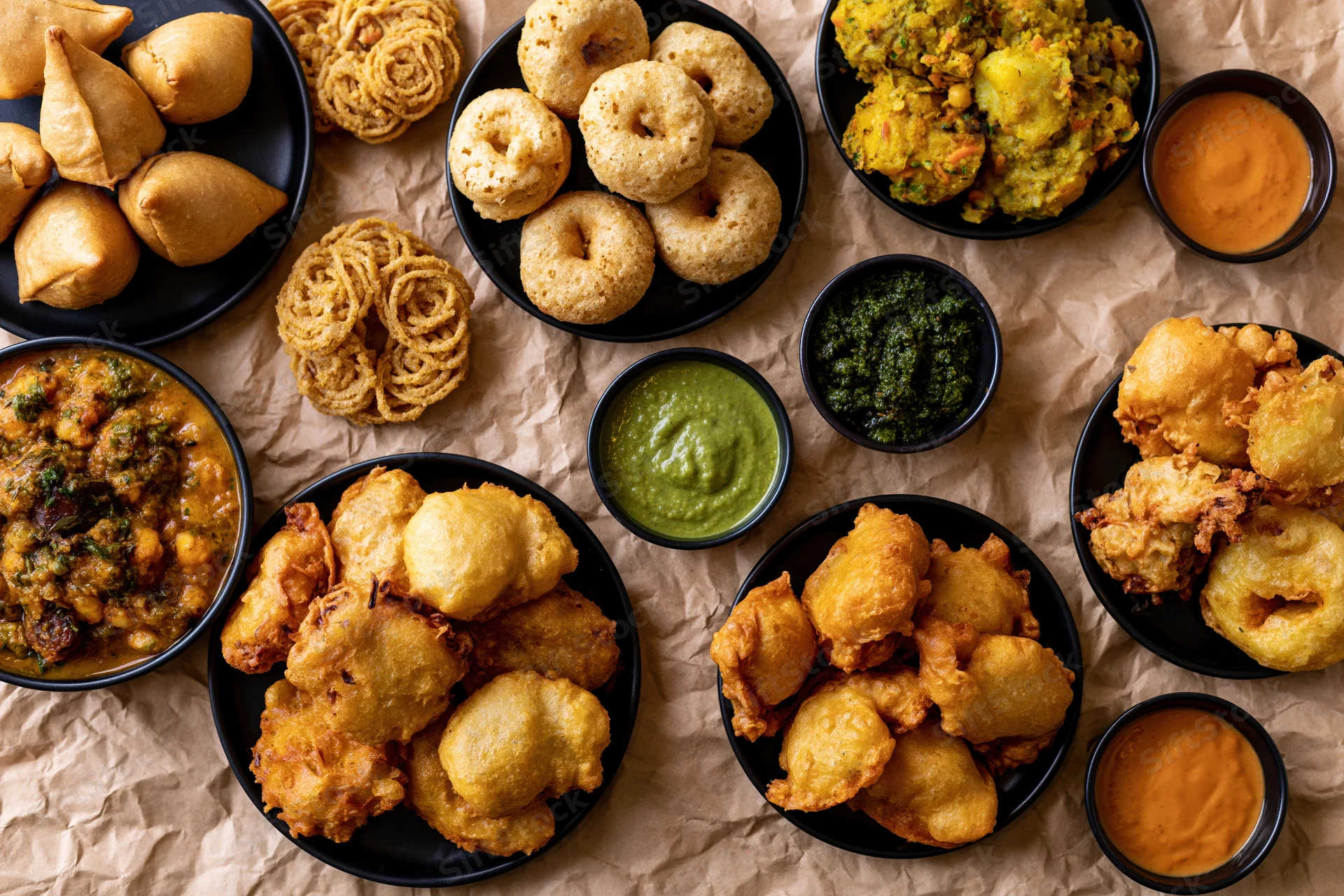 Overhead flat-lay of indian street food snacks including samosas and chutneys on crumpled brown paper