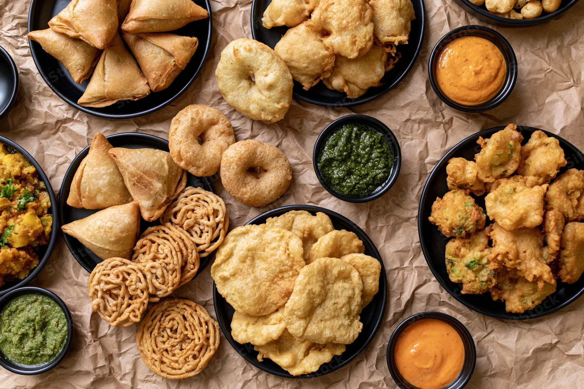 Overhead flat-lay of indian street food snacks including samosas, pakoras, and chutneys arranged on crumpled brown paper