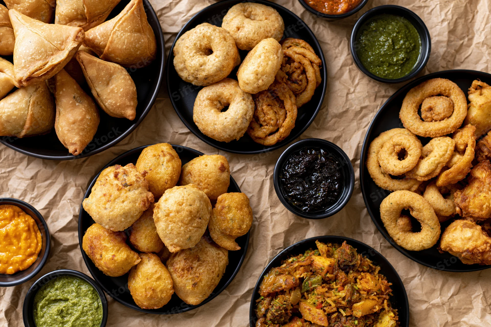 Overhead flat-lay of indian street food snacks including samosas, kachori, and onion rings with chutneys on brown paper