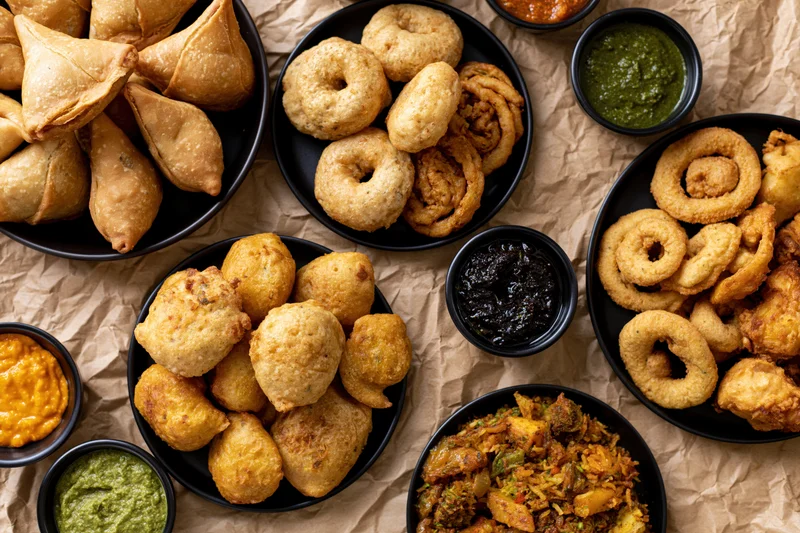 Overhead flat-lay of indian street food snacks including samosas, kachori, and onion rings with chutneys on brown paper