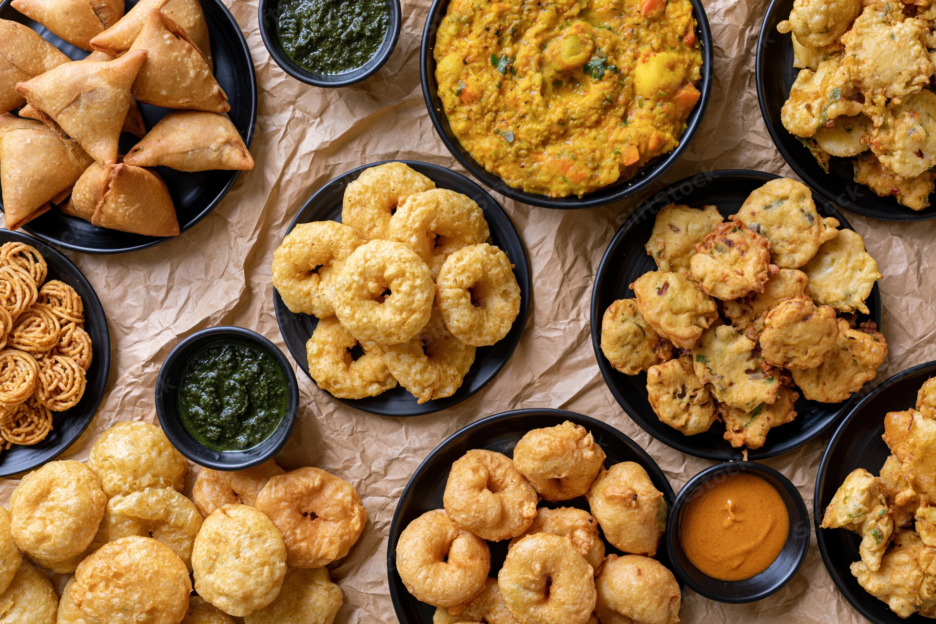 Overhead flat-lay of indian street food snacks including samosas, kachori, pakora, and chutney on black plates arranged on crumpled paper