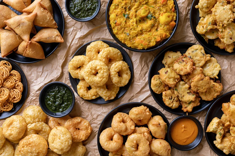 Overhead flat-lay of indian street food snacks including samosas, kachori, pakora, and chutney on black plates arranged on crumpled paper