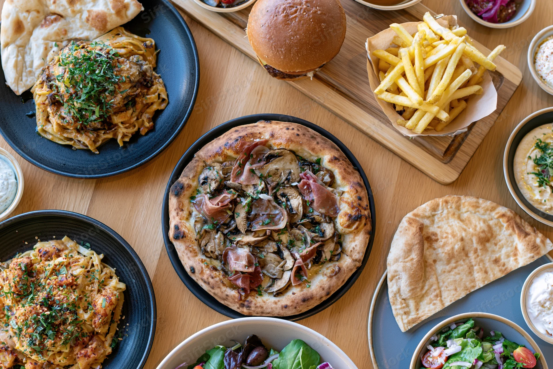 Overhead view of a restaurant table spread with pizza, burger, fries, naan, pasta, and salad on wooden surface