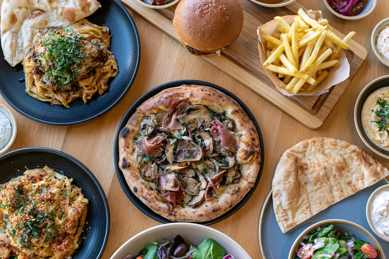 Overhead view of a restaurant table spread with pizza, burger, fries, naan, pasta, and salad on wooden surface