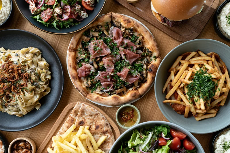 Overhead view of a restaurant table with pizza, pasta, fries, salad, burger and other dishes arranged for a shared meal